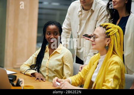 Smiling afro designer looking at her funny colleague who is pretending that she is sleeping. close up side view photo. woman hipnotising her friend, y Stock Photo