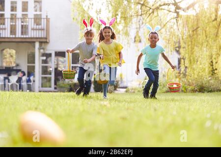Group Of Children Wearing Bunny Ears Running On Easter Egg Hunt In Garden Stock Photo