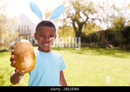 Portrait Of Boy Wearing Bunny Ears Holding Chocolate Egg On Easter Egg Hunt In Garden Stock Photo