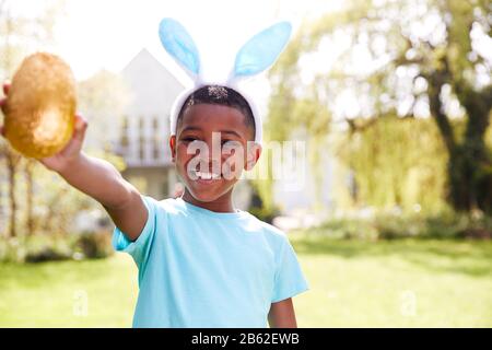 Portrait Of Boy Wearing Bunny Ears Holding Chocolate Egg On Easter Egg Hunt In Garden Stock Photo