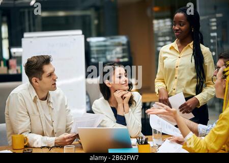 diverse people are talking in the office, young woman leaning on her palms , crossing her fingers and listening to her colleagues' opinions on the dea Stock Photo