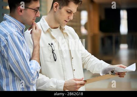 young man in glasses tocuhing his chin wiuth a hand, trying understand the problem, two active designers, journalists making up an article, writes wri Stock Photo
