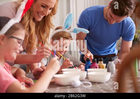 Parents With Children Wearing Bunny Ears Sitting At Table Decorating Eggs For Easter At Home Stock Photo