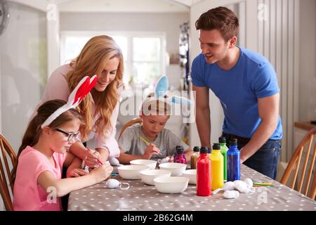 Parents With Children Wearing Bunny Ears Sitting At Table Decorating Eggs For Easter At Home Stock Photo