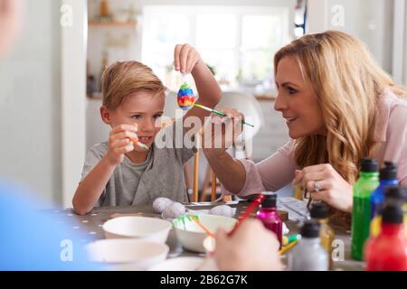 Mother With Son Sitting At Table Decorating Eggs For Easter At Home Stock Photo