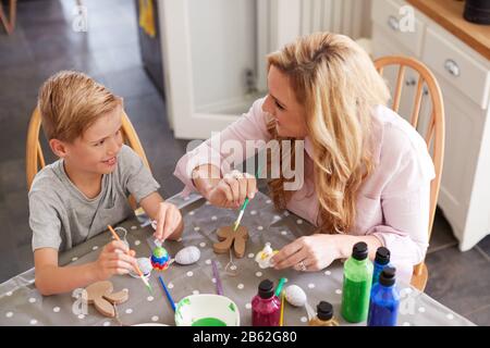 Mother With Son Sitting At Table Decorating Eggs For Easter At Home Stock Photo