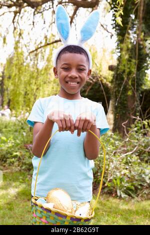 Portrait Of Boy Wearing Bunny Ears Holding Chocolate Egg On Easter Egg Hunt In Garden Stock Photo