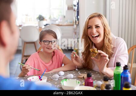 Parents With Daughter Sitting At Table Decorating Eggs For Easter At Home Stock Photo