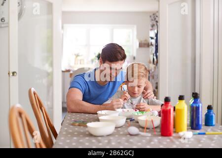 Father With Son Sitting At Table Decorating Eggs For Easter At Home Stock Photo