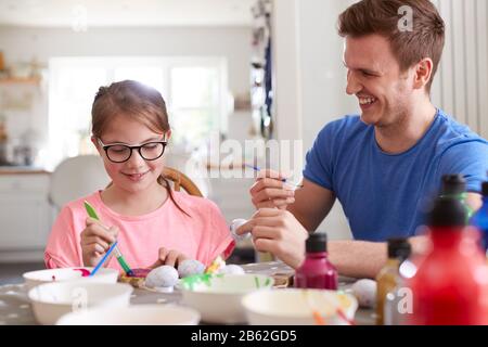 Father With Daughter Sitting At Table Decorating Eggs For Easter At Home Stock Photo