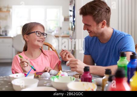 Father With Daughter Sitting At Table Decorating Eggs For Easter At Home Stock Photo