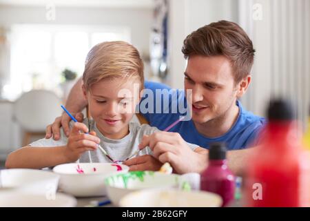 Father With Son Sitting At Table Decorating Eggs For Easter At Home Stock Photo