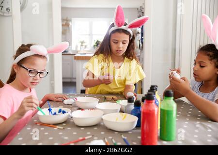 Three Girls Wearing Bunny Ears Sitting At Table Decorating Eggs For Easter At Home Stock Photo