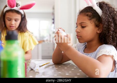 Two Girls Wearing Bunny Ears Sitting At Table Decorating Eggs For Easter At Home Stock Photo