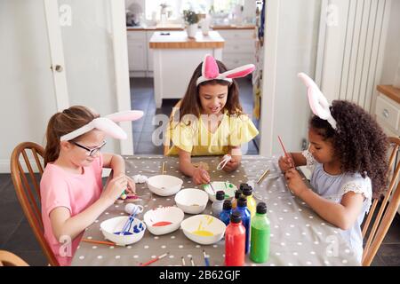 Three Girls Wearing Bunny Ears Sitting At Table Decorating Eggs For Easter At Home Stock Photo