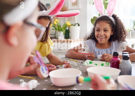 Three Girls Wearing Bunny Ears Sitting At Table Decorating Eggs For Easter At Home Stock Photo