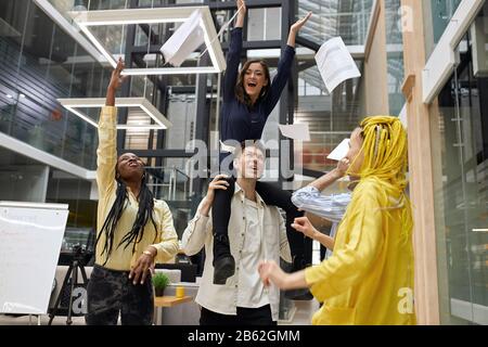 female boss sitting on her colleague and screaming, throwing paper in the air, well done, we've done it, super, hooray. crazy working day, unforgettab Stock Photo