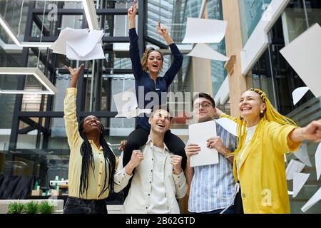 excited overjoyed young epople with raised arms throwing paper, dancing in the office. winning, best day,achievement Stock Photo