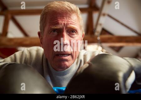 Senior Male Boxer In Gym Leaning On Ropes Of Boxing Ring Suffering From Heart Pain Stock Photo