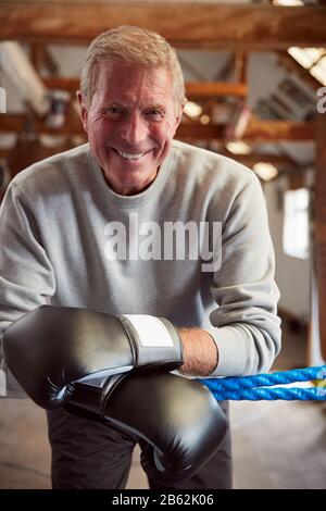 Smiling Senior Male Boxer In Gym Wearing Boxing Gloves Leaning On Ropes Of Boxing Ring Stock Photo