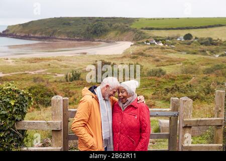 Loving Active Senior Couple Resting By Gate As They Walk Along Coastal Path In Winter Stock Photo