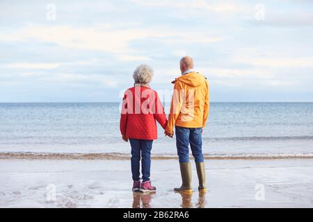 Rear View Of Senior Couple Holding Hands Looking Out To Sea On Winter Beach Vacation Stock Photo