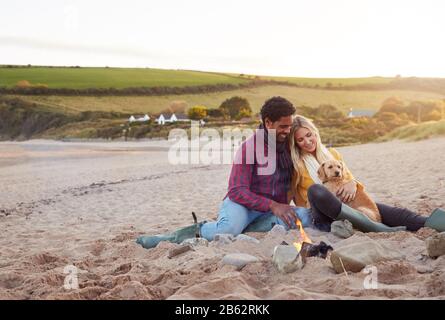 Loving Couple With Dog Relaxing By Fire On Winter Beach Vacation Stock Photo