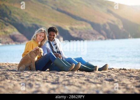 Loving Couple Sitting On Sand As They Walk With Dog Along Shoreline On Winter Beach Vacation Stock Photo
