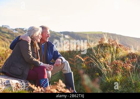 Loving Active Senior Couple Walking Along Coastal Path In Autumn Resting On Rock Together Stock Photo