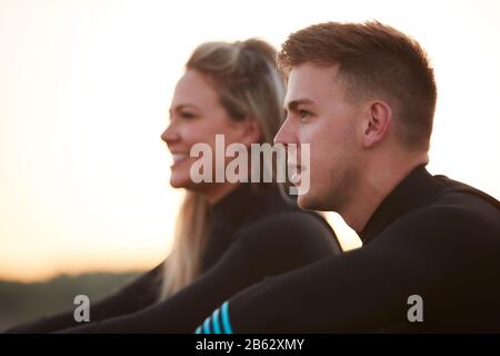 Profile View Of Couple Wearing Wetsuits On Surfing Staycation Sitting On Beach Looking Out To  Sea Stock Photo