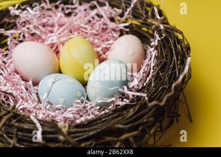 A nest of twigs and hay and a pink paper filler with pastel colored Easter eggs. Colored eggs for the holiday of light Easter. Easter card on a yellow background. Stock Photo