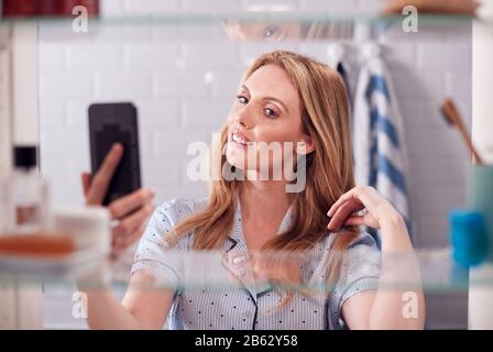 View Through Bathroom Cabinet Of Young Woman Wearing Pajamas Taking Selfie On Mobile Phone Stock Photo