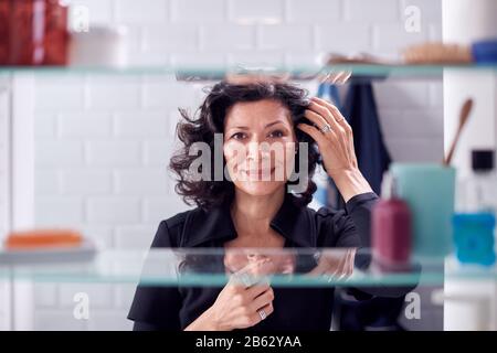View Through Bathroom Cabinet Of Mature Businesswoman Getting Ready For Work Checking Hair Stock Photo