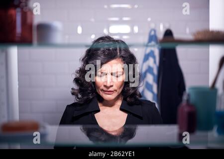 View Through Bathroom Cabinet Of Unhappy Mature Businesswoman Getting Ready For Work Stock Photo