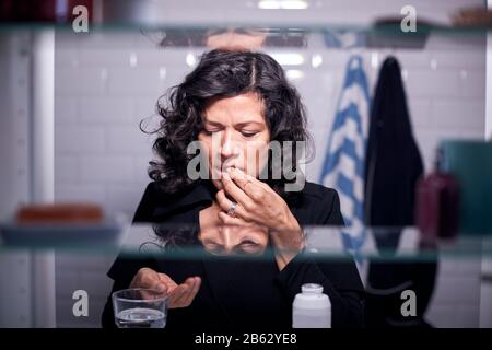 View Through Bathroom Cabinet Of Mature Woman Taking Medication With Glass Of Water Stock Photo