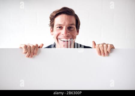 Portrait Of Happy Young Man Holding Blank White Card With Copy Space Standing Against White Wall Stock Photo