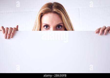 Young Woman Holding Blank White Card With Copy Space In Front Of Face Standing Against White Wall Stock Photo