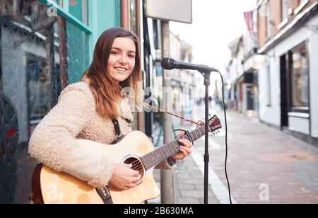 Portrait Of Young Female Musician Busking Playing Acoustic Guitar And Singing Outdoors In Street Stock Photo