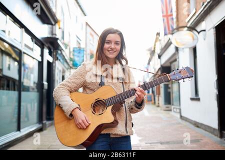 Portrait Of Young Female Musician Busking Playing Acoustic Guitar And Singing Outdoors In Street Stock Photo