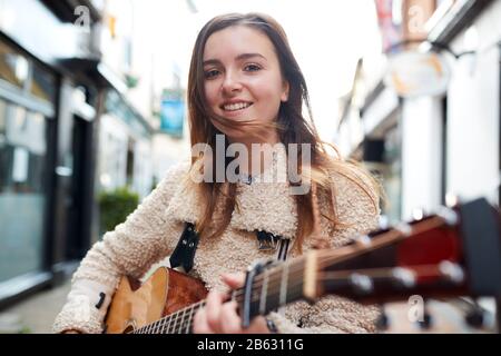 Portrait Of Young Female Musician Busking Playing Acoustic Guitar And Singing Outdoors In Street Stock Photo