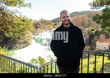 Short haired man standing on a fence on Krka "Skradinski buk" river waterfall. Smiling happily on a warm winter day in a black coat Stock Photo