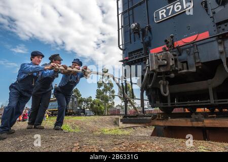 Three young male volunteers dressed in 19th century rail clothes pushing a massive steam locomotive engine on a turn bridge on a sunny day. Stock Photo