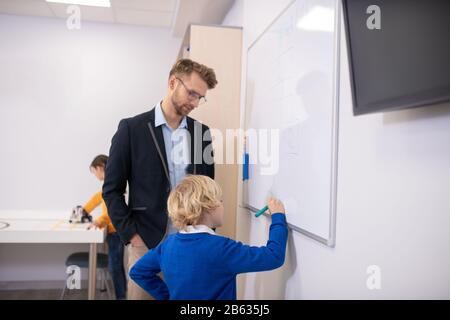 Male teacher standing at whiteboard, boy writing Stock Photo