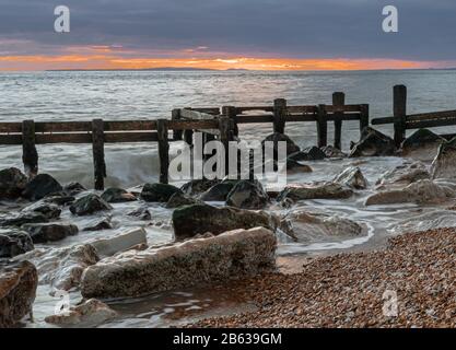 Winter seascape vista of old, battered and broken sea defences with warm glowing sunset on the distant horizon Stock Photo