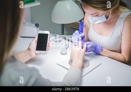 Woman in a nail salon receiving a manicure by a beautician. Smartphone in hands of client Stock Photo