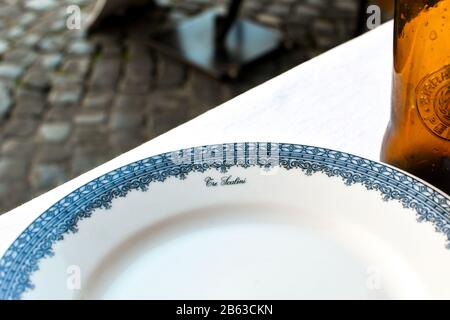 A decorated china plate from a historic restaurant in Rome Italy alongside a bottle of beer, at an outdoor cafe. Stock Photo