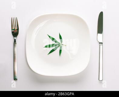 Conceptual image of a green cannabis leaf on a white plate with a knife and fork isolated on a white background with copy space for text. Don't take d Stock Photo