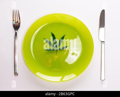 Conceptual image of green cannabis leaf on a light green plate with a knife and fork isolated on white background with copy space for text. Say no to Stock Photo