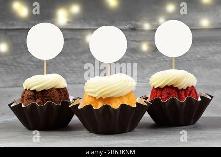 This cupcake topper mock up features three delicious gourmet cupcakes on a weathered gray wood background. Golden lights shine in the background of th Stock Photo