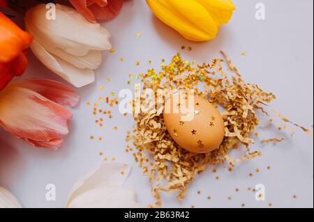 Brown chicken egg decorated with gold sequins and stars in pressed cut paper on a delicate pink background, a bouquet of multi-colored tulips in the b Stock Photo